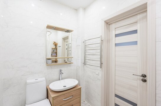 bright bathroom features white marble tiles, a wooden vanity with an oval sink, a toilet, a mirror with shelves, a heated towel rack, and a wooden door with blue accents