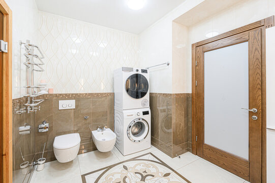 well-lit bathroom featuring a toilet, bidet, stacked washer and dryer, and a frosted glass door. The room has patterned tiles and neutral color tones