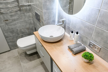 .Modern bathroom with gray tiles, white sink, wooden countertop, and round mirror with LED...