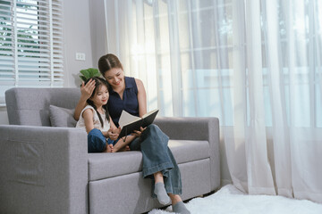 Fototapeta premium An Asian mother and daughter sit together indoors, smiling and bonding over a book, enjoying digital learning and joyful family time.