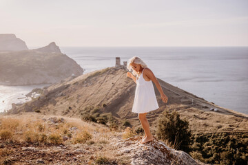 A blonde woman stands on a hill overlooking the ocean. She is wearing a white dress and she is...