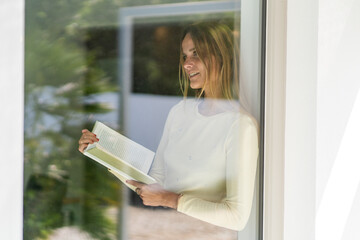 Woman by the window at home, holding a book in her hand. Woman looking thoughtful and happy, having...