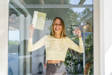 Woman by the window at home, with glass of water and a book on her hand. Woman looking happy,...
