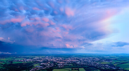 Panoramic view of vibrant mammatus clouds at sunset over a city and green fields. Ideal for: weather background, dramatic skies, nature prints, and meteorological visual content.