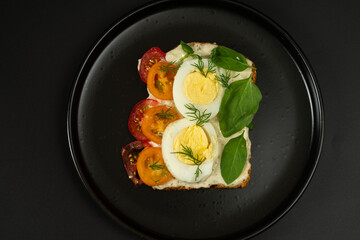 Diet sandwich on a black plate. Sandwich with boiled eggs, tomatoes and greens. Contrast of colors.