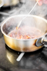 A steaming stainless steel pan with meat cooking on a stovetop. A hand stirs the ingredients with a spoon, showcasing active food preparation in a kitchen setting
