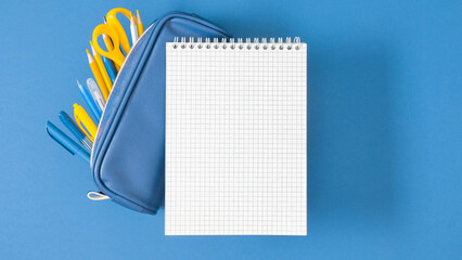 Top view of empty notebook and full pencil case of different pencils and pens as well as scissors on blue background. Back to school.