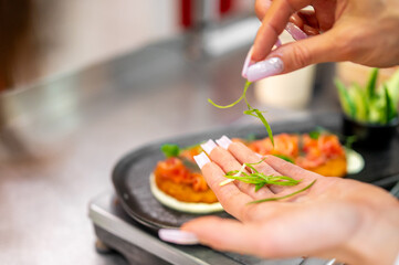 A close-up of a hand with manicured nails delicately placing fresh green herbs on a gourmet dish, showcasing culinary preparation, food styling, and fine dining aesthetics.