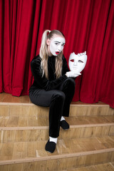 A mime girl with a mask sits on a theater stage.