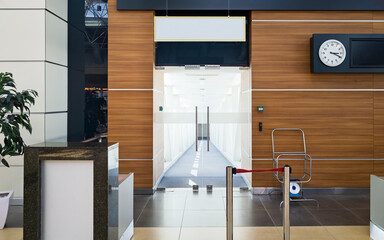 Glass doors open to empty jet bridge inside bright airport terminal. Clean floors, wooden wall panels with clock and flight information display, organized airport space for boarding passengers