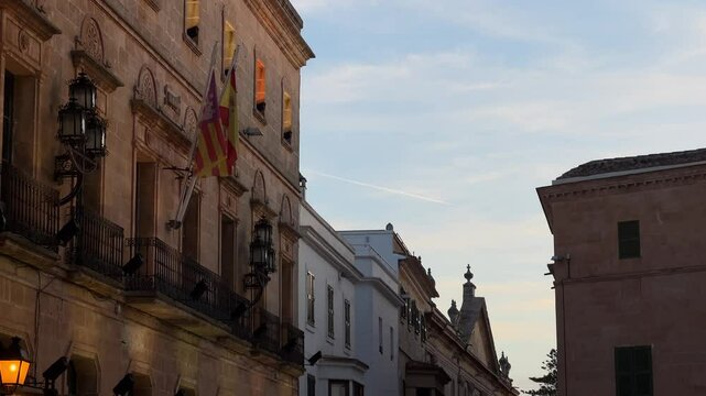 Stunning shot of Parroquia de Nuestra Se&ntilde;ora del Roser in Ciutadella, Menorca with the warm evening light highlighting its historic facade and beautiful balconies, accompanied by the regional flags.