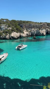 Sailboats anchored in the crystal-clear turquoise waters of Cala Macarella, Menorca, surrounded by limestone cliffs and lush Mediterranean vegetation on a sunny summer day.