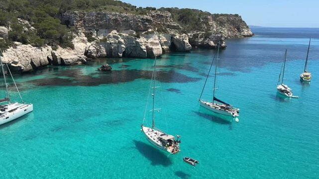 Sailboats anchored in the crystal-clear turquoise waters of Cala Macarella, Menorca, surrounded by limestone cliffs and lush Mediterranean vegetation on a sunny summer day.