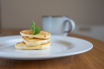 Three small pancakes garnished with mint leaf on white plate on wooden table, blurred cup in the background