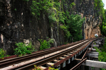 Obraz premium Train railway track between hellfire pass mountain and Si Sawat or Khwae Kwai river with forest jungle wildness in Sai Yok National Park for travel visit at Tham krasae cave in Kanchanaburi, Thailand