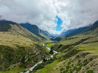Naklejka premium An aerial view shows a lush green mountain valley with a winding river flowing through it, under a dramatic cloudy sky with a patch of blue
