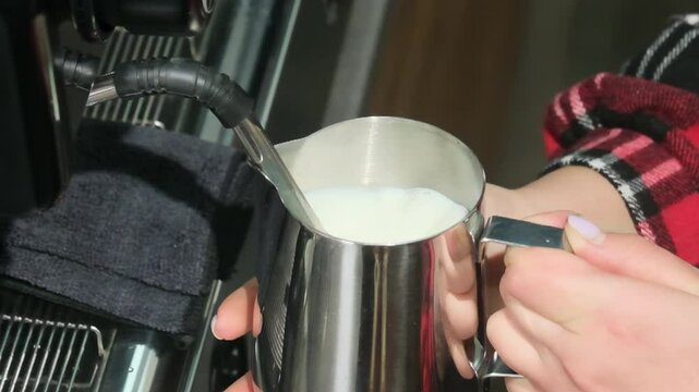 Woman barista creates frothy milk using espresso machine wand. Hand guides pitcher beneath steaming nozzle as hot vapor slow motion