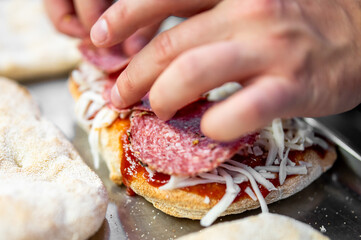 A close-up of hands making a pizza, placing slices of salami on tomato sauce and shredded cheese over fresh dough. The scene captures the homemade pizza-making process in detail.