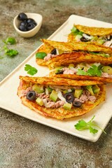 Vegetable tacos with zucchini, carrot and potato tortilla filled with canned tuna, sliced ​​avocado and olives on ceramic plate on green concrete background.