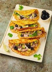 Vegetable tacos with zucchini, carrot and potato tortilla filled with canned tuna, sliced ​​avocado and olives on ceramic plate on green concrete background.