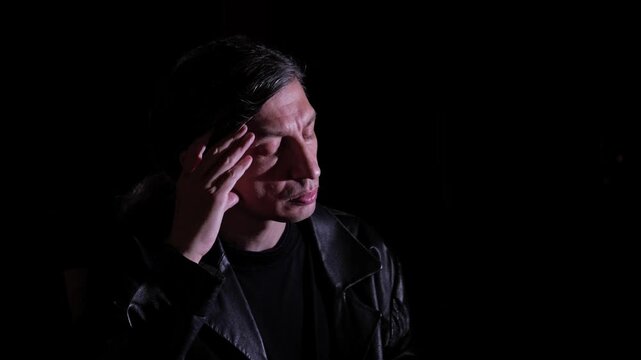 A close-up shot captures an overwhelmed and distressed mood as a man in a leather jacket covers his face with his hand, eyes closed, against a dark background, suggesting stress or despair