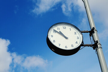 Round Outdoor Street Clock With Black Hands And Numerals Mounted On A Metal Pole Against A Bright...