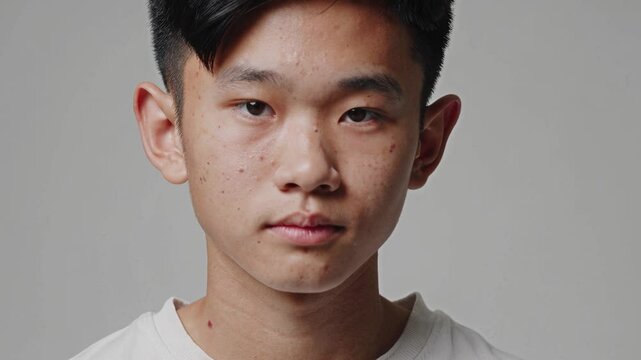 Close-up portrait of an Asian teenage boy with imperfect skin, looking confidently and naturally at the camera. A sincere and relatable image that highlights real beauty, adolescence, individuality