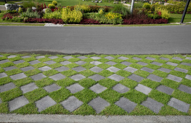 Checkered pattern of green grass and cement tiled floor with walkway and flower garden in background