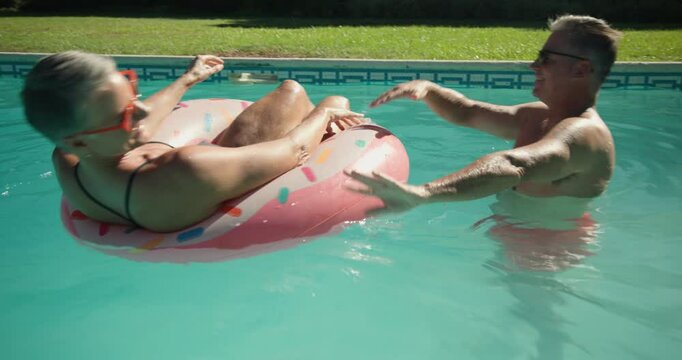 With man giving gentle push, senior couple spinning on pink donut float in backyard pool laughing