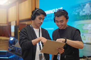Event manager discussing rundown with stage manager while pointing at clipboard, showing behind the...
