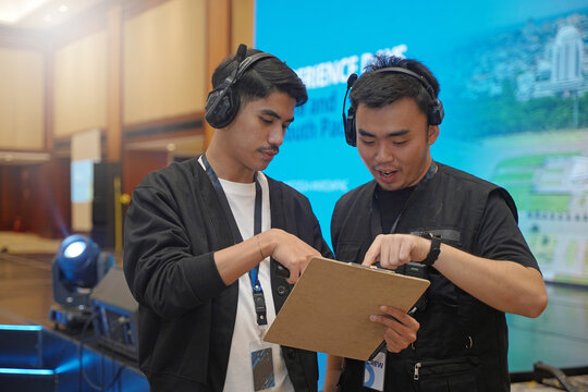 Asian male stage manager is discussing while pointing at clipboard, using headset and ID card, showing the coordination of work behind the scenes of the event with the stage atmosphere and LED screen.