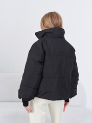 Woman wearing a black puffer jacket faces away, showing off the back detailing. Her hair is blond and loose. She stands indoors  white sofa and wall provide a minimalistic setting © Dmitry Tsvetkov