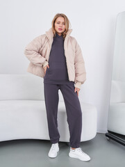 A woman with blond hair in a beige puffer jacket over a dark hoodie and matching joggers, poses...