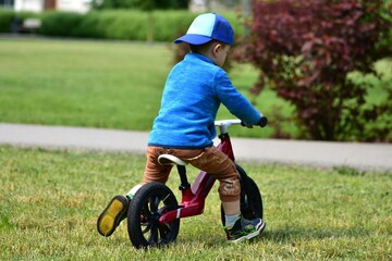 Young toddler boy riding a balance bike on a park grass. Child wearing cap and sporty outfit enjoys...