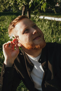 Portrait of a young happy red-haired bearded man with a white flower behind his ear on a summer day in the park.