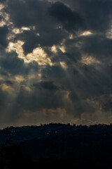 Obraz premium Crepuscular rays pierce through dark, dramatic clouds, illuminating a silhouette of distant hills.