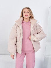 Woman in a light puffer jacket over a pink hoodie and matching pants. She stands against a mirror,...