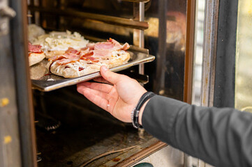 A person places a tray of uncooked pizzas into an oven. The pizzas are topped with cheese and...