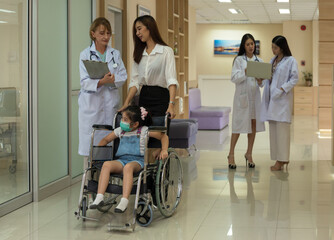 Child in wheelchair wearing face mask accompanied by mother and female doctor in hospital corridor. Two women doctors discussing medical notes in background.