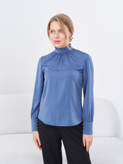A woman with blonde hair, in a blue long-sleeve blouse with a high collar, stands against a white...
