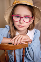 A charming young girl of European appearance in a blue dress with a white lace collar sits with her hands on the back of a chair. The child is wearing a straw hat and glasses with bright red frames.
