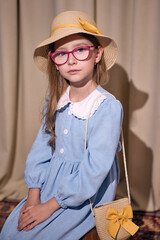A thoughtful young girl in a blue dress with a white collar sits against a light curtain. She wears a straw hat with a ribbon and pink glasses. She has a small bag with a yellow bow on her shoulder.