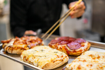 A chef uses tongs to serve freshly baked pizzas with various toppings, including pepperoni, bacon, and cheese. The close-up highlights textures, melted cheese, and crispy crust