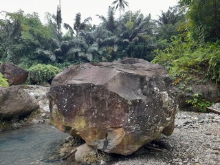 A large rock rests beside a clear stream surrounded by dense tropical vegetation in Lombok,...