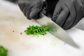 close-up of a person wearing black gloves finely chopping fresh green herbs with a sharp knife on a white cutting board. The image highlights precision, cleanliness, and professional food preparation