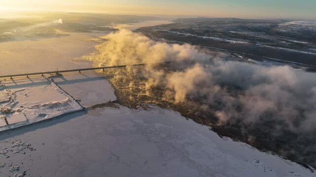 Aerial view of a wide frozen river with mist rising from it, a bridge, and snow-covered banks, in the Murmansk region, Russia