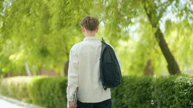 Back view of schoolboy walking with backpack and laptop in schoolyard
