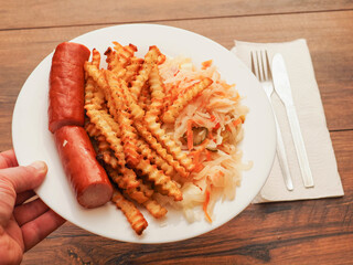 A plate of food with a hot dog and French fries on it and sauerkraut salad. The plate is on a wooden table. Tasty dinner.