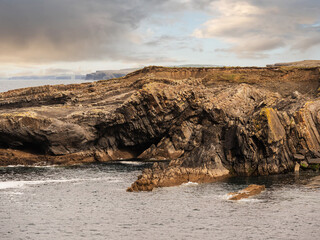 A rocky cliff coastline by the ocean. The water is calm and the sky is overcast. The scene is peaceful and serene. Bridges of Ross area, Ireland. Cloudy sky. Popular tourist landmark with amazing view