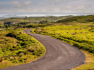 Small s shaped road with green fields on a side and lake with many small islands under cloudy sky in the background. Stunning nature scenery in Connemara, Ireland. Travel and tourism.